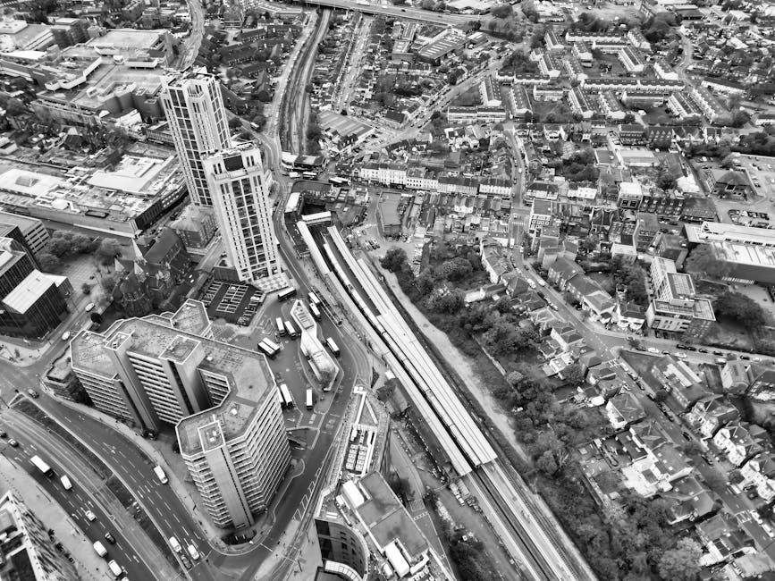 An aerial black-and-white photograph of a dense urban area showing a mix of high-rise and mid-rise buildings, residential houses, and tree-lined streets. A railway track runs diagonally across the image, with a train station platform visible alongside it. The streets are filled with vehicles, including buses and cars, and some buses are parked near the bus stops. The photograph captures a busy city environment typical for London, near Harringay station, illustrating an active transportation hub and surrounding residential and commercial areas. The image is part of a visual overview of urban infrastructure, related to house removals and furniture transport services offered by Man and Van Harringay, with the scene emphasizing the complexity of moving logistics in a city setting.