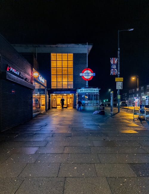 This nighttime street view shows the entrance to Harringay Green Lanes station, with illuminated signs including the London Underground roundel and a nearby coffee shop called ZIE Coffee & Co. The station entrance is framed by a glass facade with warm orange lighting inside, contrasting with the dark sky. To the right, a lamppost and a few narrow buildings with lit windows are visible along the street, which is wet and reflects the surrounding lights. The pavement in front of the station has scattered boxes and small plastic-wrapped items, suggesting preparations for a home relocation or moving process. A person is seen near the station entrance, and a man and van service like Man and Van Harringay may be involved in the moving tasks indicated by the scene. The overall environment illustrates an urban setting prepared for moving activities, with elements consistent with furniture transport, packing, and loading operations typically associated with house removals near Harringay Green Lanes station.