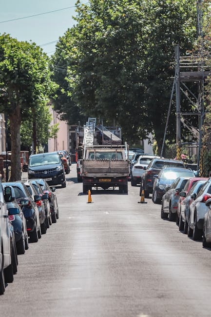 A narrow residential street with parked cars lining both sides, including sedans and hatchbacks, and tall leafy trees overhead providing shade. In the middle of the street, a flatbed truck is in the p
