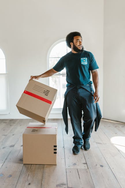 A man with dark skin and an afro hairstyle is standing inside a bright, spacious room with large arched windows, holding a cardboard box wrapped with red packing tape in his right hand. He is dressed