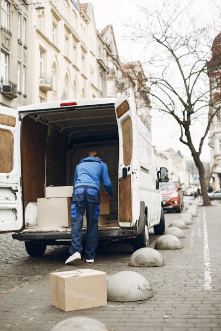 A man wearing a blue jacket and dark blue trousers is loading large, wrapped furniture pieces into the back of a white cargo van parked on a city street. The open rear doors reveal the wooden interior of the van, which is being used for furniture transport as part of a home relocation process. Several cardboard boxes, some on the pavement, are nearby, indicating packing materials used for moving. The street is lined with old, white multi-story buildings with ornate facades, and leafless trees suggest a cold or winter season. In the background, parked cars and a row of large, rounded stone bollards along the curb are visible, with natural daylight illuminating the scene. This activity aligns with the logistics of house removals and professional moving services, as performed by companies like Man and Van Harringay.