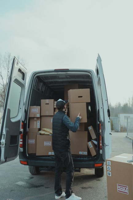 A man wearing a dark jacket, jeans, and a cap is standing inside the open back of a white moving van, which is parked outdoors on a paved surface. The van's interior is filled with stacked cardboard boxes of various sizes, some sealed with packing tape and labeled with barcode stickers, indicating a home relocation process. The man is lifting or positioning a large box, suggesting he is involved in the loading or unloading of household items. Additional boxes are visible on the ground near the van, with one marked as fragile. The sky above is overcast, and the environment appears to be a suburban or industrial area, with some trees and structures in the background. The scene reflects professional packing and furniture transport activities typical of a moving service, such as those provided by Man and Van Harringay, supporting efficient and careful home removal logistics.