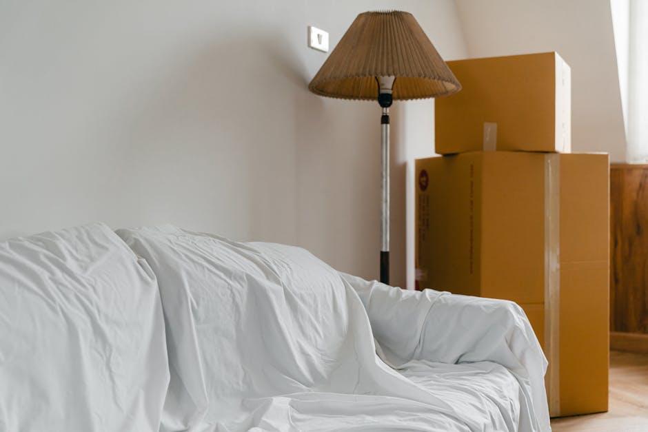 Photo of a living room corner showing a white sofa covered with a protective sheet, positioned near a beige wall. Behind the sofa, there is a tall, standing floor lamp with a brown lampshade. To the right of the sofa, three large cardboard boxes, of varying sizes, are stacked on top of each other and placed on a wooden floor. The boxes appear to be sealed and are likely used for packing belongings during home relocation. The scene is well-lit, with natural light coming from a source outside the frame. In the context of house removals and furniture transport, this image depicts the packing and loading process, with items prepared for safe transportation by Man and Van Harringay, possibly in preparation for a large furniture move involving access considerations.