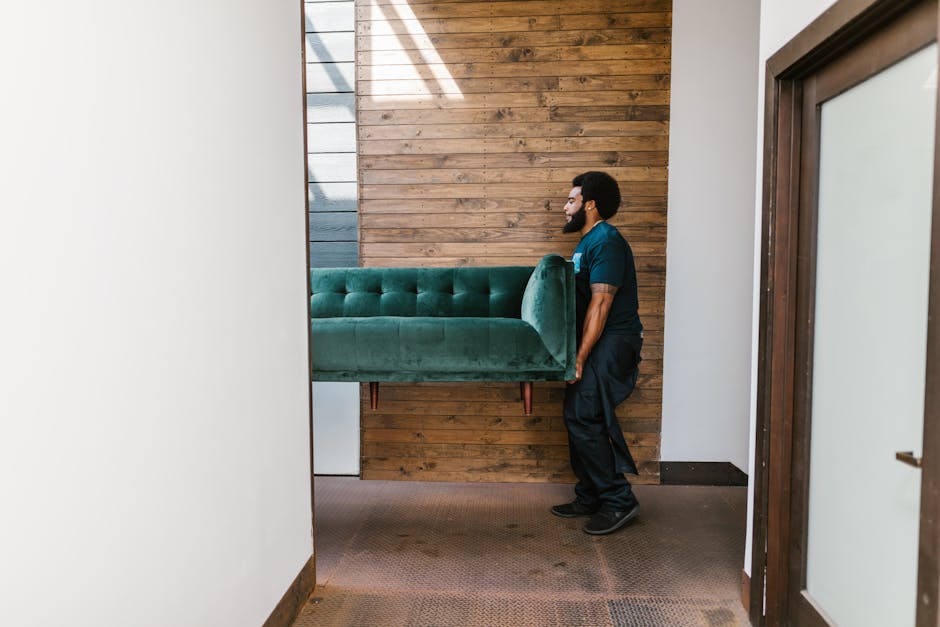 A man wearing a teal uniform is lifting a green upholstered sofa with wooden legs, manoeuvring it through a doorway inside a property. The room features a wooden accent wall behind him and white walls on either side, with a brown carpeted floor. Sunlight streams through a window or skylight, casting light on the scene. This process is part of a home relocation or furniture transport operation, with the sofa being carefully moved to the next location. The workspace appears organized, with no visible packaging materials or additional furniture in the immediate area. The image exemplifies the careful handling and logistical coordination involved in professional removals, as provided by Man and Van Harringay, to overcome access problems during large furniture removals for house moves.