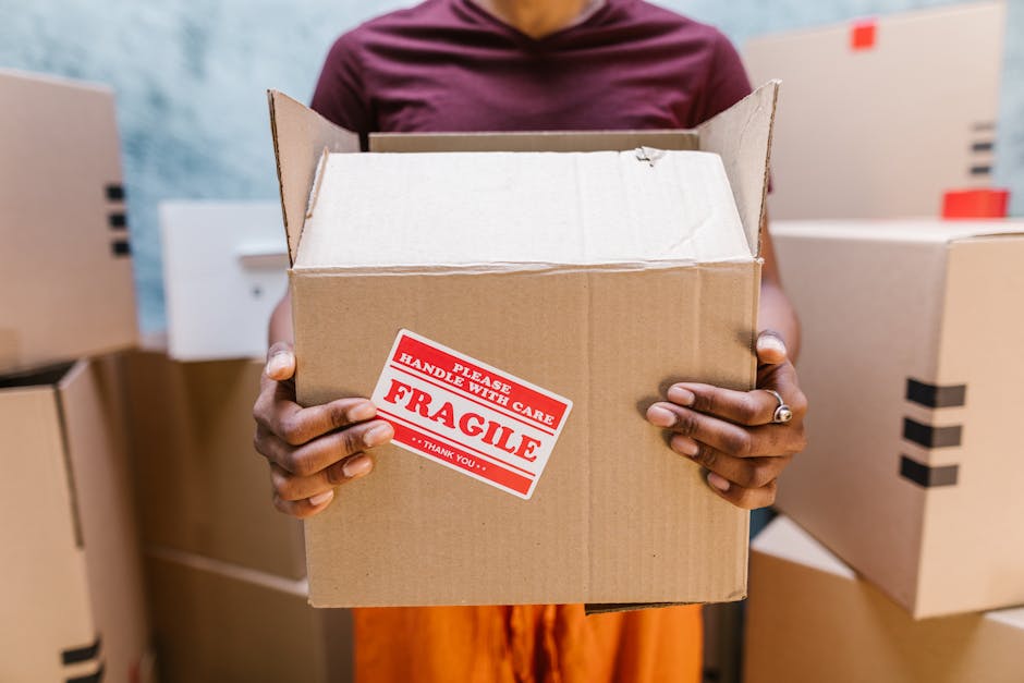 A person holding an open, medium-sized cardboard box with both hands, displaying a red and white 'Fragile' handling sticker on the front. The individual is standing in an indoor space with several other cardboard boxes of various sizes stacked or placed nearby, some sealed with black tape. The background includes a blurred wall and part of a doorway or opening, suggesting a home or storage environment during a home relocation or moving process. The person’s face is not visible, focusing on the box and the careful handling of packaging materials used in furniture transport and packing for house removals, as facilitated by Man and Van Harringay.