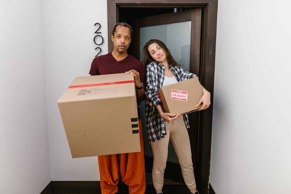 A man and a woman are standing inside a property near the doorway, each holding cardboard boxes for home relocation. The man, dressed in an orange pair of trousers and a maroon t-shirt, is holding a large, sealed cardboard box with a shipping label on the side, while the woman, wearing beige pants and a plaid shirt over a white top, carries a smaller box marked with a red and white 'FRAGILE' label. Behind them, the door frame is visible along with a section of a white wall, indicating the interior entrance of a house or apartment. The scene captures the process of packing and moving, with the individuals appearing prepared for furniture transport or a house removal service. The environment is well-lit with natural or artificial light, emphasizing the logistical phase of packing and loading boxes for an upcoming move, as handled by Man and Van Harringay.
