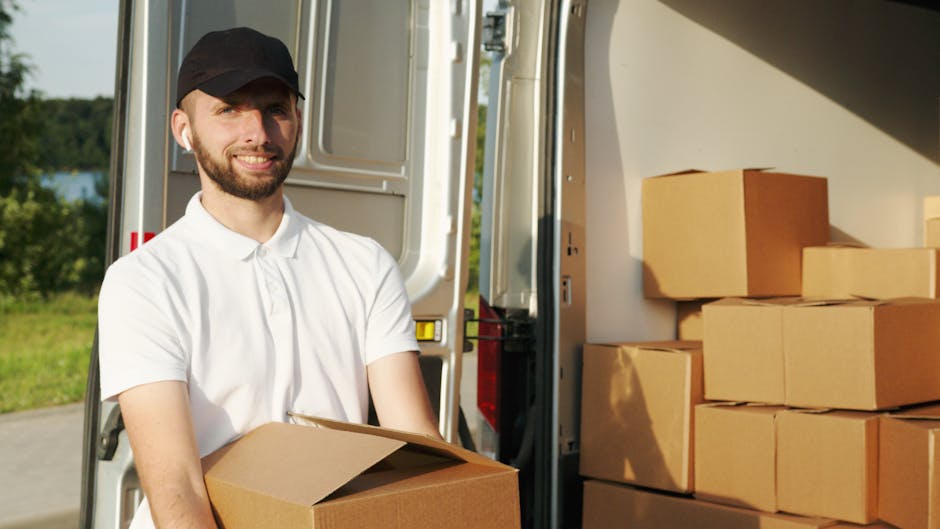 A man with a short beard, wearing a black cap and a white polo shirt, is standing outdoors beside an open moving van during daytime. He is holding a brown cardboard box, preparing to load it into the vehicle. The van's interior is visible, showing several neatly stacked cardboard boxes of various sizes, some wrapped in plastic. The area appears to be a driveway or street with a background of green trees and foliage under clear weather. The scene depicts the loading process involved in a home relocation, with the man engaged in packing or furniture transport as part of house removals services. Man and Van Harringay is providing the moving assistance, with equipment such as trolleys or straps potentially used but not visible in this shot, emphasizing the logistics of packing and moving items efficiently.
