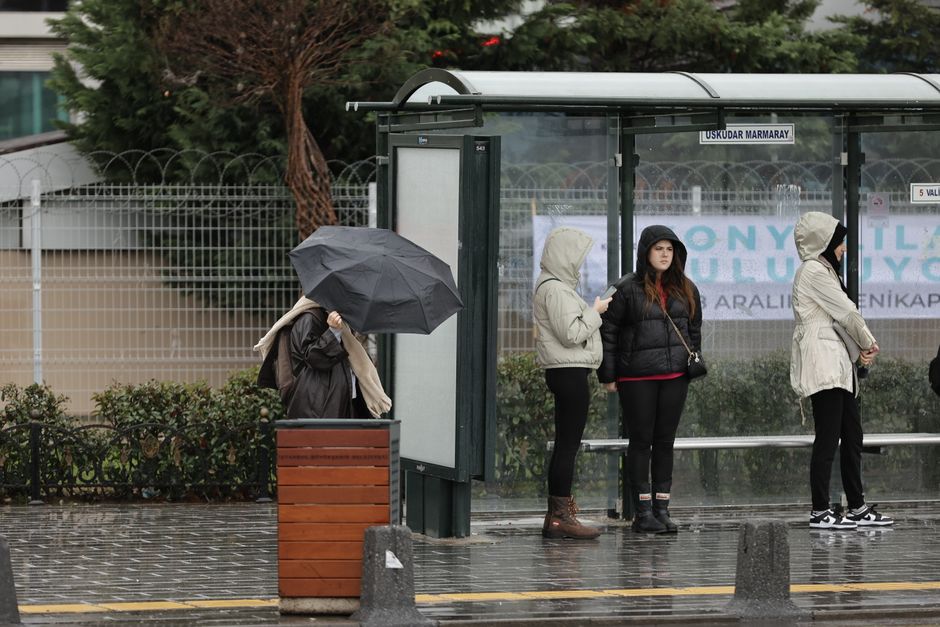 A group of four people waiting at a bus stop during rainy weather on a wet pavement, with one person holding a large black umbrella. The individuals are dressed in casual jackets, some with hoods, and are standing near a modern glass and metal shelter with informational posters and signs, including one displaying the name 'USKUDAR MARMARAY.' Behind the bus stop, there is a metal fence, some greenery, and trees, indicating an urban environment. The scene is illuminated by overcast natural light, highlighting the damp conditions, and the individuals appear to be engaged in their own activities, with some using their smartphones. This setting relates to home relocation and moving logistics, where loading and packing might occur, involving transport arrangements and outdoor waiting during a move or delivery process facilitated by a professional service such as Man and Van Harringay.