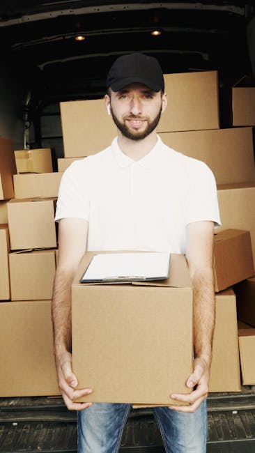 A man with a short beard, wearing a black cap and a white polo shirt, is standing outdoors beside an open moving van during daytime. He is holding a brown cardboard box, preparing to load it into the vehicle. The van's interior is visible, showing several neatly stacked cardboard boxes of various sizes, some wrapped in plastic. The area appears to be a driveway or street with a background of green trees and foliage under clear weather. The scene depicts the loading process involved in a home relocation, with the man engaged in packing or furniture transport as part of house removals services. Man and Van Harringay is providing the moving assistance, with equipment such as trolleys or straps potentially used but not visible in this shot, emphasizing the logistics of packing and moving items efficiently.